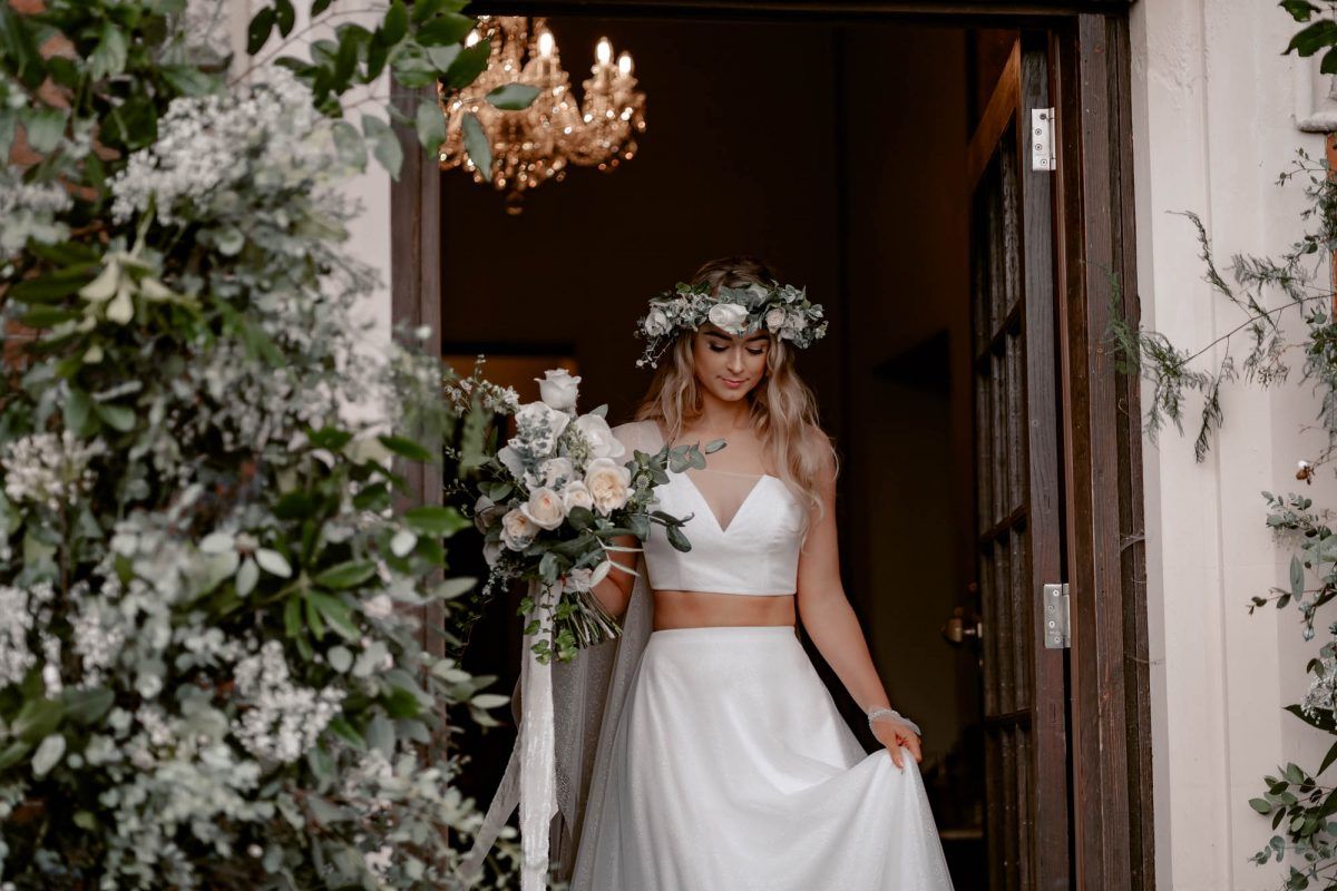A bride in a crown of flowers steps out through a doorway framed in white blossom