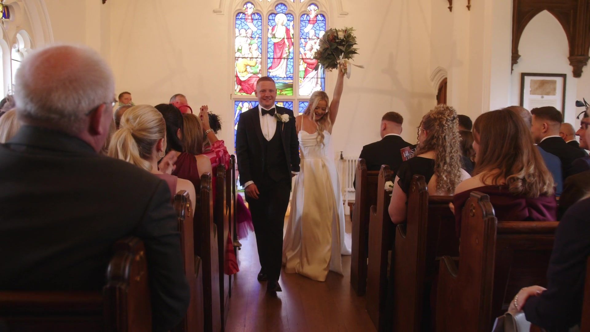 Meg raises her bouquet as she and Phil walk back down the chapel aisle beneath the stained glass