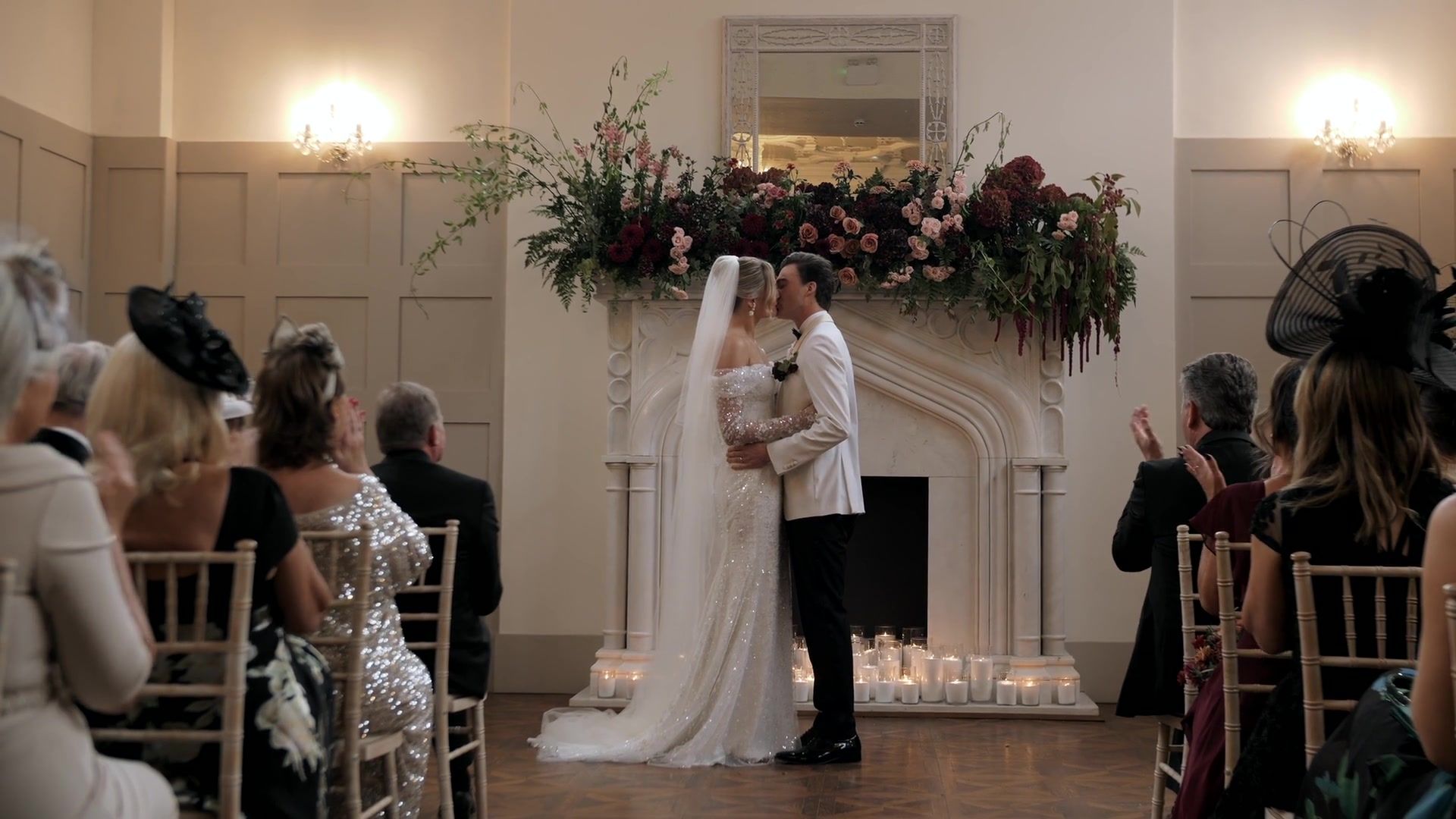 Issy and Miles kiss before the carved stone fireplace in the Morning Room as their guests applaud