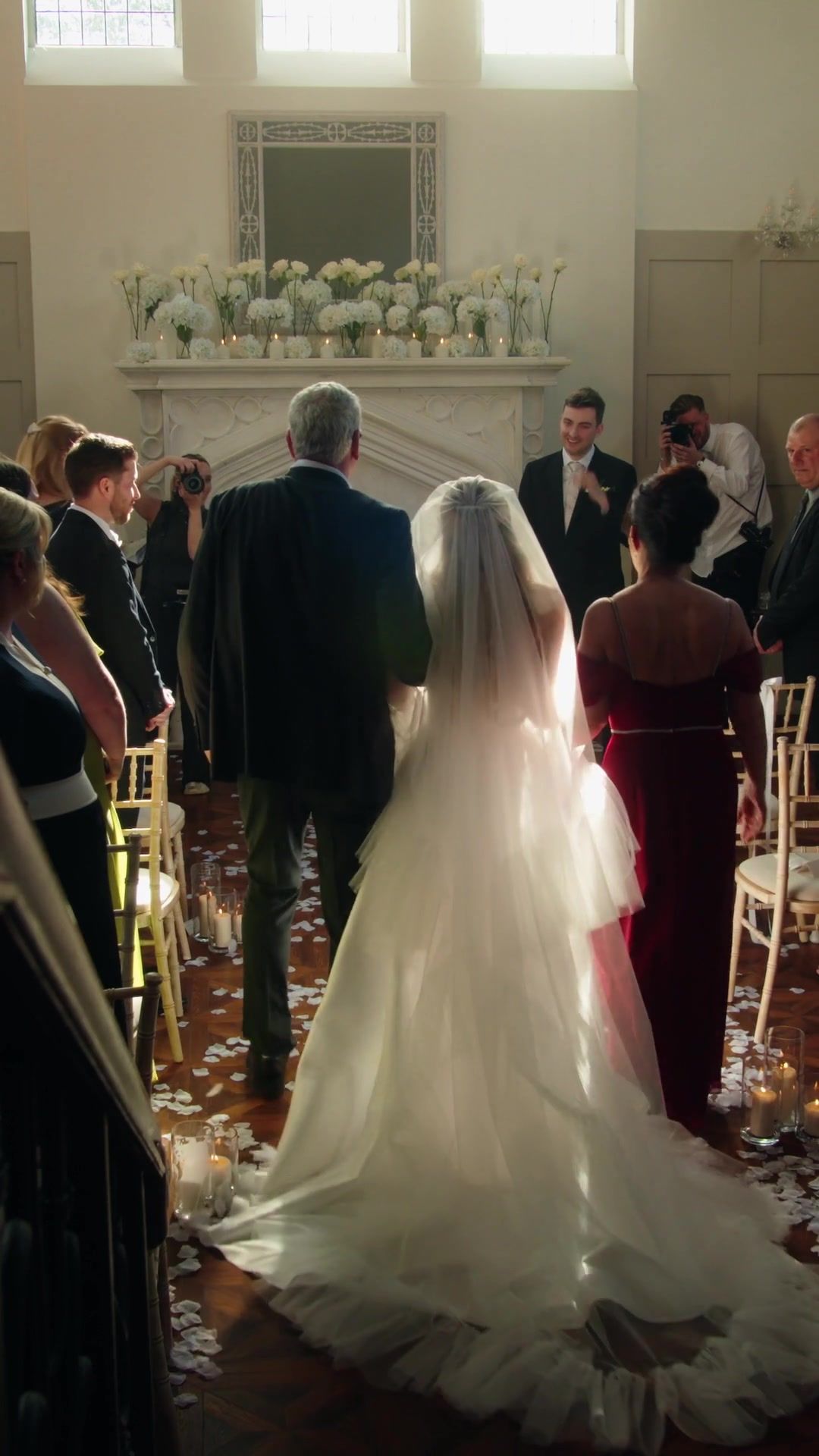 A bride walks down the aisle of the Morning Room on her father’s arm, her veil catching the light